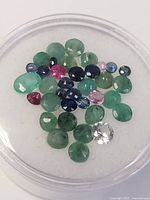 Top view of various green, blue, pink/red, and one white gemstone in a clear container on white background
