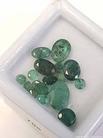 Collection of thirteen loose emeralds with varying sizes, shapes, and green hues inside a white display tray, showing natural inclusions.