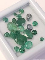 Close-up image of 19 loose emerald stones placed in a white display box showing various cuts and color depths.