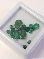 Lot of natural emerald gemstones shown in a white display box, various cuts and sizes visible.