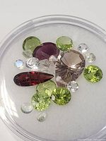 Top view of the assortment of genuine faceted gemstones including green peridot, white/blue moonstone, red garnet, purple ametrine, and smoky quartz in a round clear container.