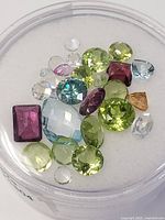 Round container holding a mix of green, purple, blue, clear, and yellow faceted gemstones of different cuts on a white background.