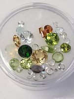 Top-down view of assorted faceted genuine gemstones with various colors including green peridot, purple amethyst, orange garnet, translucent moonstones on white padding.
