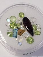 Top view image of various loose genuine gemstones including peridot, moonstone, garnet, blue topaz, and clear stones arranged in a round container.