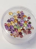 Photo showing multiple small faceted gemstones in a round transparent container, with various colors including purple, yellow, green, red, and others.