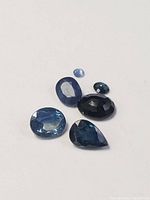 Five loose sapphire gemstones in various cuts and shades of blue on a white background, showing the size and color variation in natural sapphires.