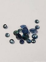 Photo showing all loose sapphire stones in various cuts and sizes with blue and greenish-blue tones, arranged on white background.