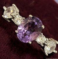 Close-up front view of silver ring with oval purple amethyst center stone flanked by round white topaz stones on a dark background.