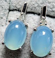 Close-up of silver earrings showing oval chalcedony cabochon stones and round diamonds on hooks, displayed on a grey textured background.