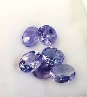 Six loose oval cut tanzanite gemstones showing facets and light to medium violet-blue shades on a white background.