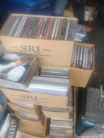 Stacked cardboard boxes labeled SRI filled with assorted CDs, some sealed cases visible