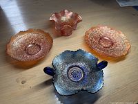 Four carnival glass pieces on wooden floor: two orange bowls, one small orange ruffled bowl, one blue handled ruffled bowl