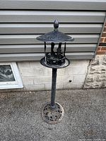 Full view of the black cast aluminum bird feeder standing against a wall on an asphalt surface, showing its height and base design.