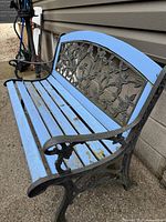 Side angle view showing cast iron armrest with floral design and blue wooden slats of bench seat and upper backrest