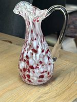 Photo of the Chalet Glass pitcher showcasing red and white spotted pattern with clear handle and ruffled rim, set on wooden surface.