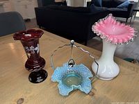 Three glass pieces displayed on wooden table: cranberry vase, blue hobnail basket with handle and spoon, and pink and white Jack in the Pulpit vase.