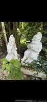 Two large concrete garden gnome statues outdoors surrounded by greenery.
