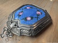 Octagonal silver-tone compact pendant with blue guilloche enamel top decorated with pink roses. Slight wear visible on metal edges and a slightly bent chain eye loop.