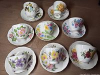 Top view of the eight English cups and saucers with various colorful floral patterns arranged in two rows on a wood floor.