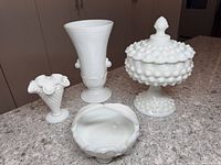 Four white milk glass hobnail items on a gray countertop including a tall covered compote dish, two vases, and a small bowl.