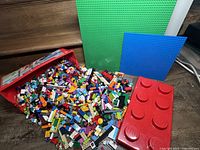 Large red Lego Juniors plastic box filled with assorted Lego pieces, with two baseplates (green and blue) and one of the two red no-name base pieces visible. Pieces spill partially outside the box.
