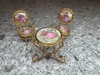 Miniature Limoges porcelain table and two chairs showing the delicate hand-painted romantic scenes on the porcelain tabletop and chair backs, with gold-tone metal frames.