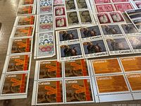Close up of blocks and strips of unused Canadian stamps including Olympic logos, historical figures, and postal maps in brown and orange tones.