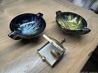 Two colorful Murano art glass ashtrays (cobalt blue and yellow) with copper accents placed on a wooden surface alongside a small metal folding travel ashtray.