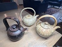 Photo showing all three pottery teapots on a wood table, with dark brown and two light cream colored pieces visible