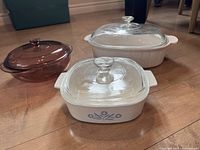 Front view of three cookware dishes: large white oval Corning Ware, smaller square white Corning Ware with blue floral pattern, and cranberry Pyrex round dish, all with lids.