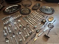 Photo showing wide assortment of silver plated spoons, forks, knives, and several serving trays and bowls on wood floor.