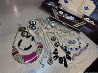 Overview photo showing multiple vintage jewellery pieces including necklaces and earrings spread out on light wood surface beside open jewellery box containing more items.