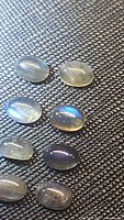 Eight oval cabochon natural labradorite gemstones laid out on a black textured surface showing their blue flash iridescence under lighting.