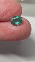 Close-up image of a natural oval cut emerald held between fingers showing size, deep green color, faceting and luster.
