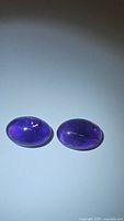 Pair of natural deep purple oval cabochon amethyst gemstones on plain background with some visible inclusions including rainbow effect in one stone.