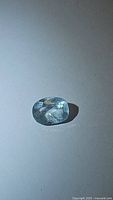 Close-up image of a cushion cut light blue aquamarine gemstone with visible inclusions, placed on a plain surface under light showing gemstone facets and color.