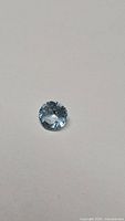 Clear photo of a single round cut blue topaz gemstone on a plain background, showing its size, color, and cut.