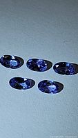 Five small oval cut tanzanite gemstones arranged on a flat surface, showing blue-purple coloration under light.