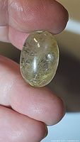 Close-up photo of natural golden rutilated quartz oval cabochon gemstone held between fingers, showing internal golden needle rutile inclusions and polished surface.