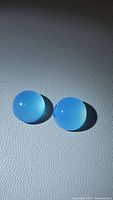 Pair of natural round cabochon icy blue chalcedony gemstones on textured gray surface, lit with cool lighting.