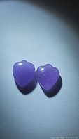 Pair of heart-shaped lavender jade stones with a purple hue, polished surfaces, and natural inclusions visible under light.