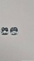 Matched pair of natural sky blue cushion cut topaz gemstones, approx 6x6 mm each, showing light blue color and clarity on white background.