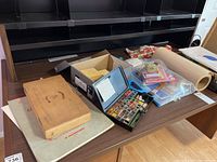 Photo showing a watercolor paint set, a cardboard box, and assorted papers including card stock and art pads on a wooden surface.