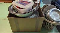 Cardboard box containing nested stainless steel mixing bowls, plastic container with cookie cutters on top, and baking tins beside box.