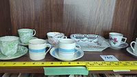 Photo showing five fine china teacups and six saucers including green floral patterned cups, white cups with blue stripes, a cup with rose detail, along with a white lattice-style serving plate.