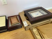 Stacks of assorted picture frames in various sizes and finishes including dark brown, light brown, reddish tones, some with backing paper inside and some with glass.