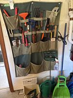 Full view of wall hanging gardening tool organizer with pockets containing various hand tools, spray bottle, and two green watering cans below.
