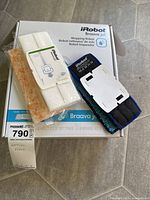 Photo of the iRobot Braava jet 240 mopping robot box with cleaning pads and accessory pad holder on top, various package contents visible.