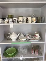 Shelf display of assorted mugs, ceramic teapots, sugar dish, plates, and clear glass tumblers with some floral designs.