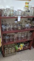 Wide shot of wooden shelf unit holding four shelves filled with various clear glass preserving jars of multiple sizes and lid types, along with boxed canning supplies at bottom shelf.
